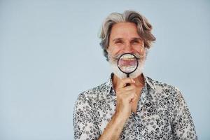 avendo divertimento con ingrandimento bicchiere. anziano elegante moderno uomo con grigio capelli e barba in casa foto