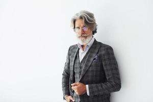 in posa per un' telecamera mentre in piedi contro bianca sfondo. anziano elegante moderno uomo con grigio capelli e barba in casa foto