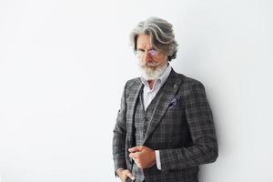 in posa per un' telecamera mentre in piedi contro bianca sfondo. anziano elegante moderno uomo con grigio capelli e barba in casa foto