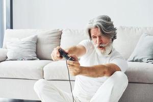 giochi video gioco di utilizzando controllore. anziano elegante moderno uomo con grigio capelli e barba in casa foto