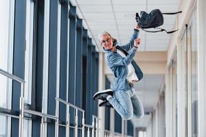 giovane maschio alunno nel occhiali, con bianca capelli e zaino è salto e avere divertimento nel corridoio di Università foto