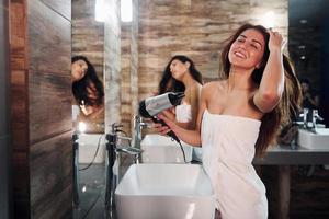 Due bellissimo giovane donne in piedi nel bagno vicino il specchio insieme, pulizia loro facce e essiccazione capelli foto