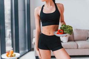 detiene salutare cibo nel mani. giovane donna con sottile corpo forma nel nero abbigliamento sportivo avere fitness giorno in casa a casa foto