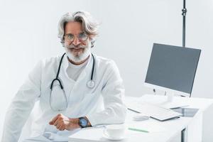 lavori di computer tavolo. anziano maschio medico con grigio capelli e barba nel bianca cappotto è in casa nel clinica foto