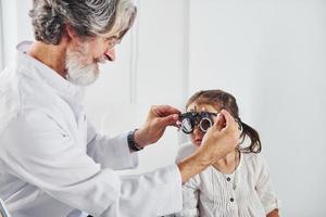 anziano maschio oculista con grigio capelli e barba nel bianca cappotto è in casa nel clinica analisi vista di poco ragazza foto
