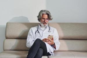 anziano maschio medico con grigio capelli e barba nel bianca cappotto è in casa nel clinica si siede su divano con Telefono nel mani foto