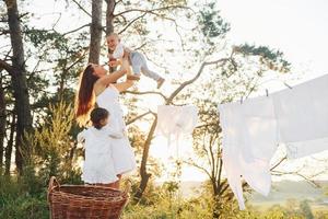 bianca Abiti sospeso su il corda per asciutto. giovane madre con sua poco figlia e figlio è all'aperto nel il foresta. bellissimo luce del sole foto