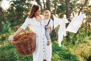 positivo casalinga con si blocca bianca Abiti per asciutto. giovane madre con sua poco figlio è all'aperto nel il foresta. bellissimo luce del sole foto