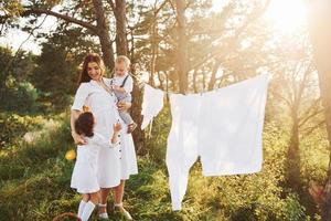 bianca Abiti sospeso su il corda per asciutto. giovane madre con sua poco figlia e figlio è all'aperto nel il foresta. bellissimo luce del sole foto