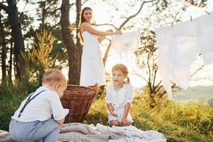 bianca Abiti sospeso su il corda per asciutto. giovane madre con sua poco figlia e figlio è all'aperto nel il foresta. bellissimo luce del sole foto