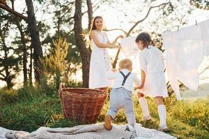 bianca Abiti sospeso su il corda per asciutto. giovane madre con sua poco figlia e figlio è all'aperto nel il foresta. bellissimo luce del sole foto