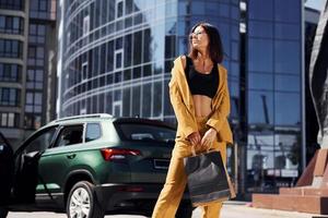 moderno attività commerciale edificio a sfondo. giovane alla moda donna nel Borgogna colorato cappotto a giorno con sua auto foto