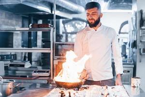 frittura padella è su fuoco. capocuoco nel bianca uniforme cucinando cibo a cucina. occupato giorno a opera foto