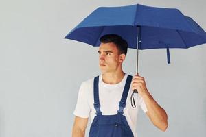 detiene ombrello di mano. maschio lavoratore nel blu uniforme in piedi dentro di studio contro bianca sfondo foto