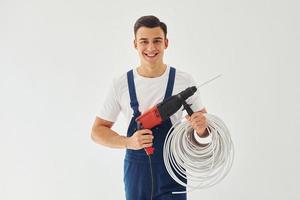 con trapano e cavi nel mani. maschio lavoratore nel blu uniforme in piedi dentro di studio contro bianca sfondo foto