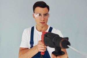 nel protettivo occhiali e con trapano nel mani. maschio lavoratore nel blu uniforme in piedi dentro di studio contro bianca sfondo foto