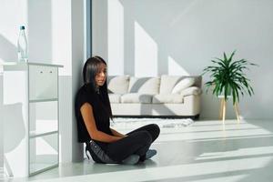 giovane brunetta nel nero Abiti si siede su il pavimento a casa a giorno foto