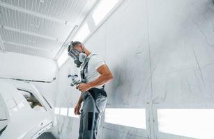 con pittura pistola. caucasico automobile riparatore nel uniforme lavori nel box auto foto