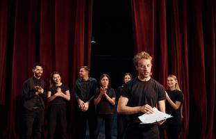 uomo con bloc notes pratica il suo ruolo. gruppo di attori nel buio colorato Abiti su prova nel il Teatro foto