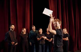 uomo con bloc notes pratica il suo ruolo. gruppo di attori nel buio colorato Abiti su prova nel il Teatro foto