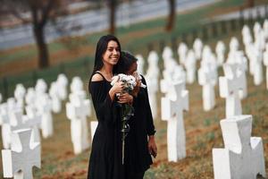 ragazza sorridente per no Motivo. Due giovane donne nel nero Abiti visitare cimitero con molti bianca croci. concezione di funerale e Morte foto