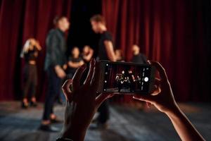 donna di mani detiene Telefono e assunzione immagine. gruppo di attori nel buio colorato Abiti su prova nel il Teatro foto