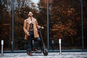 contro edificio e con elettrico scooter. giovane maschio modello nel alla moda Abiti è all'aperto nel il città a giorno foto