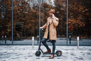 su il elettrico scooter. giovane maschio modello nel alla moda Abiti è all'aperto nel il città a giorno foto