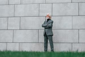 anziano uomo d'affari nel formale Abiti, con grigio capelli e barba è all'aperto sta contro il parete foto