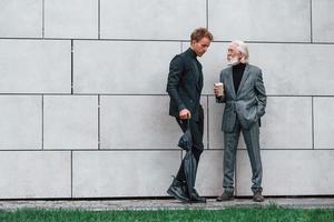 ombrello e tazza di bere. giovane tipo con anziano uomo nel elegante Abiti è all'aperto insieme. concezione di attività commerciale foto