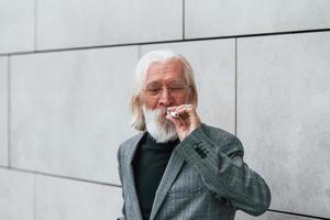 anziano uomo d'affari nel formale Abiti, con grigio capelli e barba è all'aperto fuma elettronico sigaretta foto