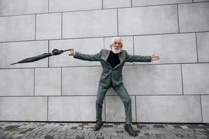 detiene ombrello. anziano uomo d'affari nel formale Abiti, con grigio capelli e barba è all'aperto foto