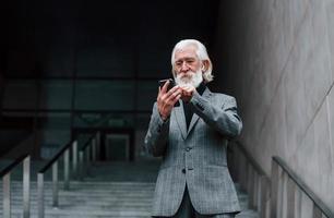 anziano uomo d'affari nel formale Abiti, con grigio capelli e barba è all'aperto provando senza fili cuffie con Telefono foto