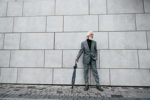 detiene ombrello. anziano uomo d'affari nel formale Abiti, con grigio capelli e barba è all'aperto foto
