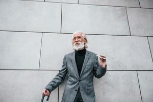 anziano uomo d'affari nel formale Abiti, con grigio capelli e barba è all'aperto fuma elettronico sigaretta foto