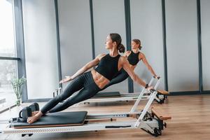 in piedi su Palestra attrezzatura e fare si allunga. Due donne nel allegro indossare e con sottile corpi avere fitness yoga giorno in casa insieme foto