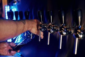 uomo versare alcool nel il bicchiere. vicino su Visualizza di birra rubinetti nel un' pub. artificiale blu illuminazione foto