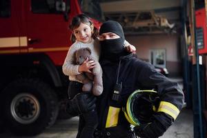 detiene orsacchiotto orso. contento poco ragazza è con maschio pompiere nel protettivo uniforme foto