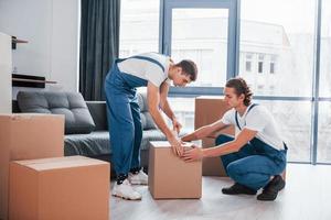 confezione il scatola. Due giovane traslocatori nel blu uniforme Lavorando in casa nel il camera foto