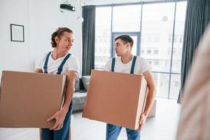 occupato giorno. Due giovane traslocatori nel blu uniforme Lavorando in casa nel il camera foto