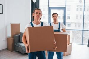 con scatole nel mani. Due giovane traslocatori nel blu uniforme Lavorando in casa nel il camera foto