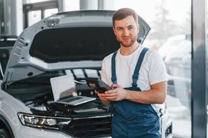 usi il computer portatile. giovane uomo nel bianca camicia e blu uniforme riparazione automobile foto