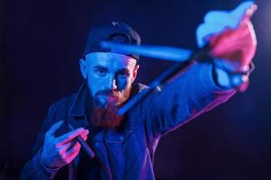 con forbici. giovane barbuto barbiere nel berretto in piedi nel il studio con neon illuminazione foto