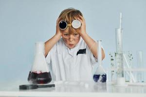 impaurito poco ragazzo nel cappotto giocando un' scienziato nel laboratorio di utilizzando attrezzatura foto