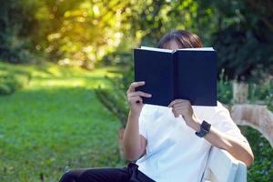 asiatico donna Tenere un' libro nel davanti di sua viso mentre seduta e lettura parco panchina angolo. concetto. asiatico donna fare all'aperto attività, come come lettura libri, Lavorando, avendo un' picnic con famiglia foto