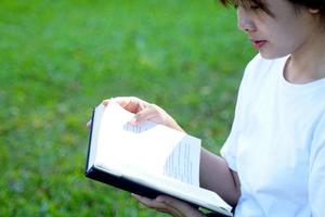 asiatico donna seduta e lettura un' libro su il erba nel il parco con concetto fare all'aperto attività come come lettura, Lavorando, ascoltando per musica, avendo un' picnic, eccetera. morbido e selettivo messa a fuoco. foto