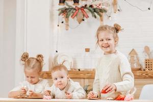 3 bionda ragazze decorare Natale biscotti con glassatura su un' di legno tavolo nel il cucina. il amiche siamo cucinando insieme nel il cucina con Natale arredamento. foto