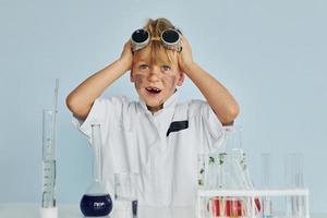 impaurito poco ragazzo nel cappotto giocando un' scienziato nel laboratorio di utilizzando attrezzatura foto