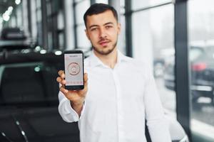 detiene Telefono con etichette e icone. giovane uomo nel bianca camicia è in casa con moderno nuovo automobile. a distanza controllo foto