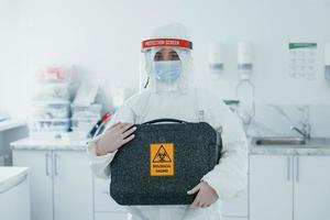 detiene Astuccio con biologico rischio. femmina medico lavoratore nel protettivo uniforme lavori su covid-19 vaccino foto